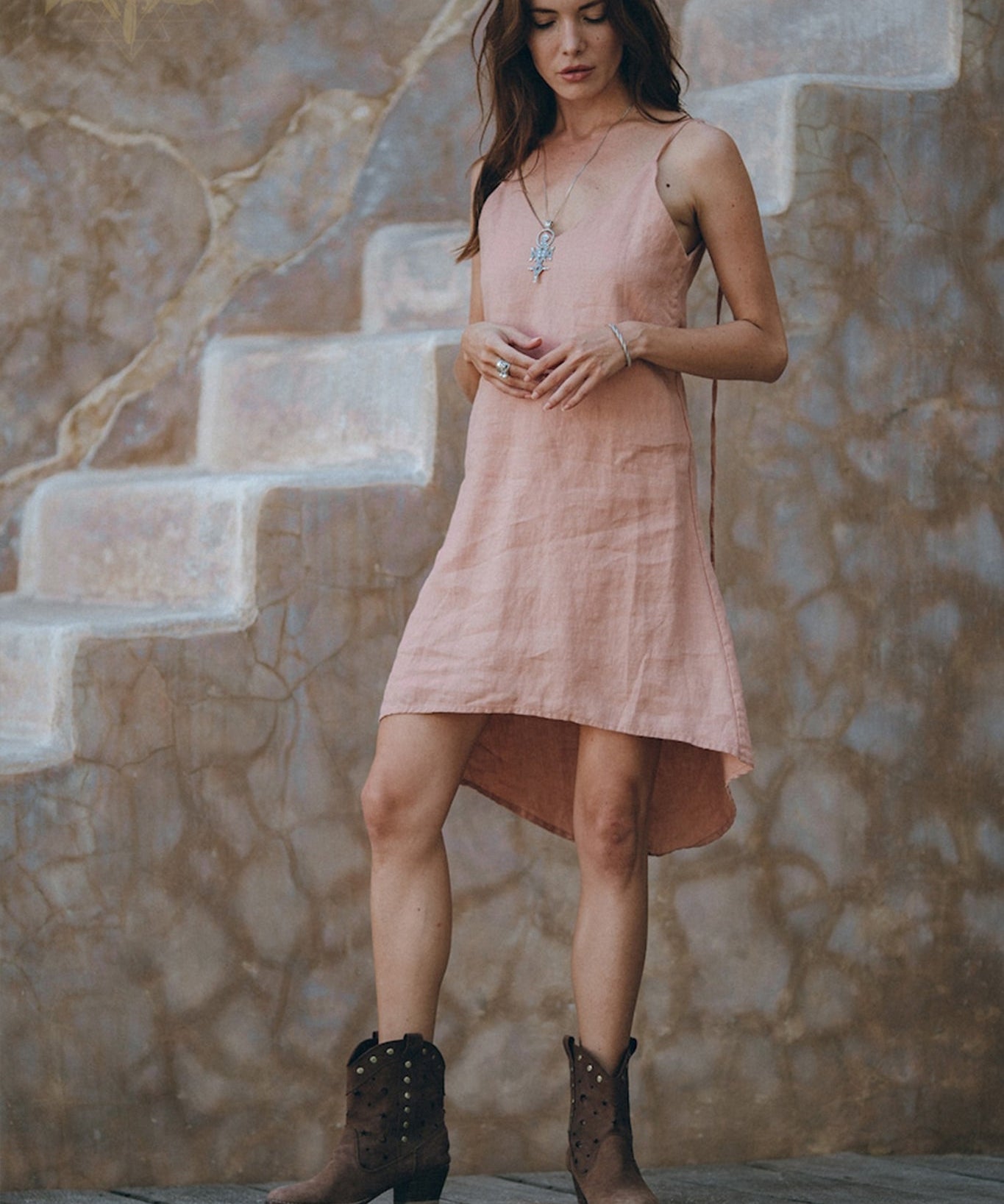 A woman wearing the AYA Sacred Wear Linen Boho Bridesmaid Dress in a pink hue pairs it with brown boots as she stands on a wooden floor. She gazes down, her hand gently touching a necklace. In the background, a rustic wall with faded stairs enhances the scene's earthy, vintage vibe with its sustainable ambiance.