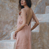 A woman with long hair wearing the AYA Sacred Wear Linen Boho Bridesmaid Dress, a pink linen bohemian slip midi dress, poses on stone steps, looking over her shoulder. The background features a textured wall with earthy tones, capturing the essence of sustainable fashion.