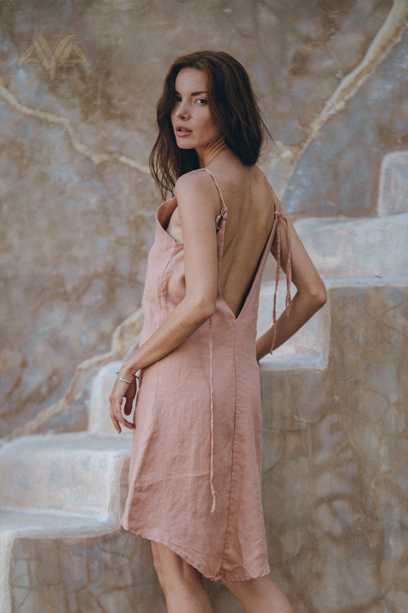 A woman with long hair wearing the AYA Sacred Wear Linen Boho Bridesmaid Dress, a pink linen bohemian slip midi dress, poses on stone steps, looking over her shoulder. The background features a textured wall with earthy tones, capturing the essence of sustainable fashion.