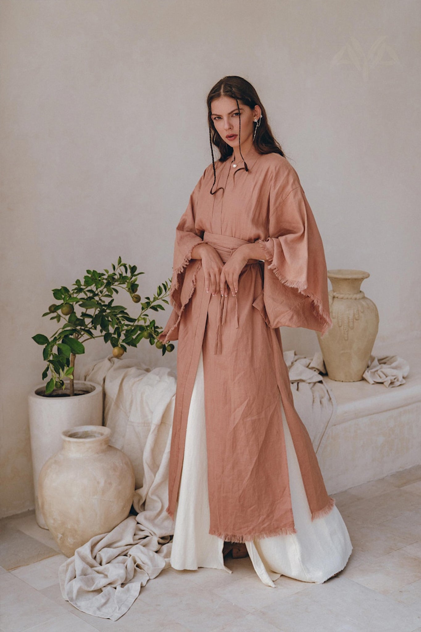 A woman stands in a minimalist setting, draped in the AYA Sacred Wear Linen Cardigan for women, featuring a dusty pink boho wrap design with a belt. Her dark hair cascades down as she gazes towards the camera, surrounded by large ceramic vases and lush green plants.