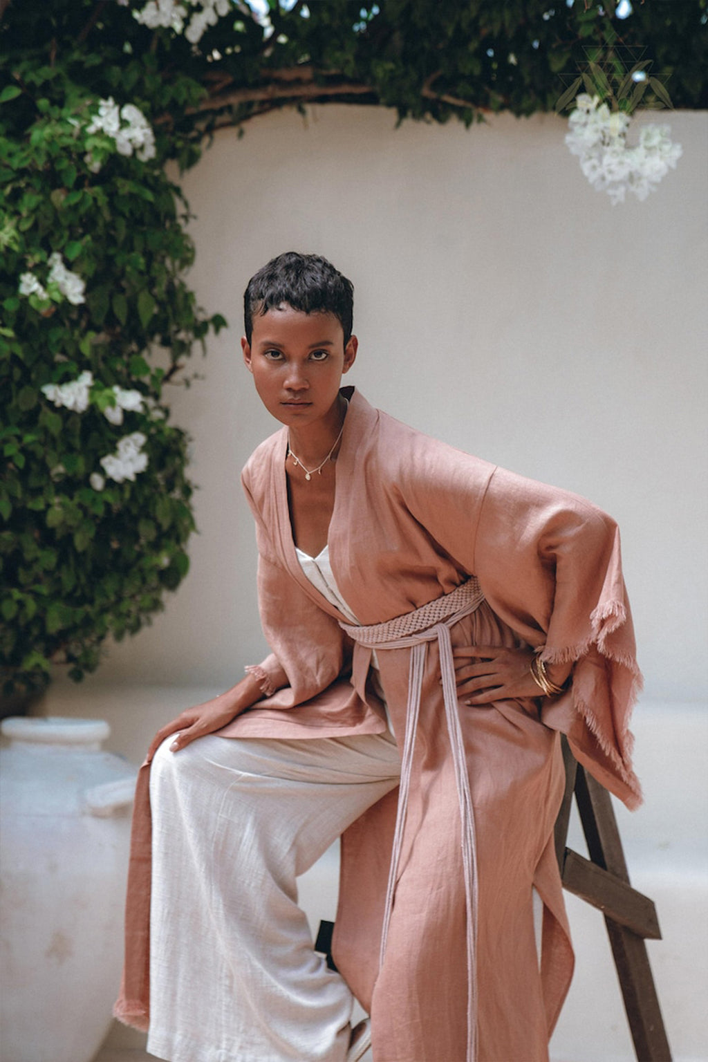 A person with short hair poses outdoors, wrapped in the AYA Sacred Wear Linen Cardigan Women, a boho wrap cardigan with a belt in dusty pink hues. They pair it elegantly with white pants as they sit gracefully on a wooden stool. The scene is enchanting, enhanced by a large pottery vase and lush greenery adorned with white flowers.