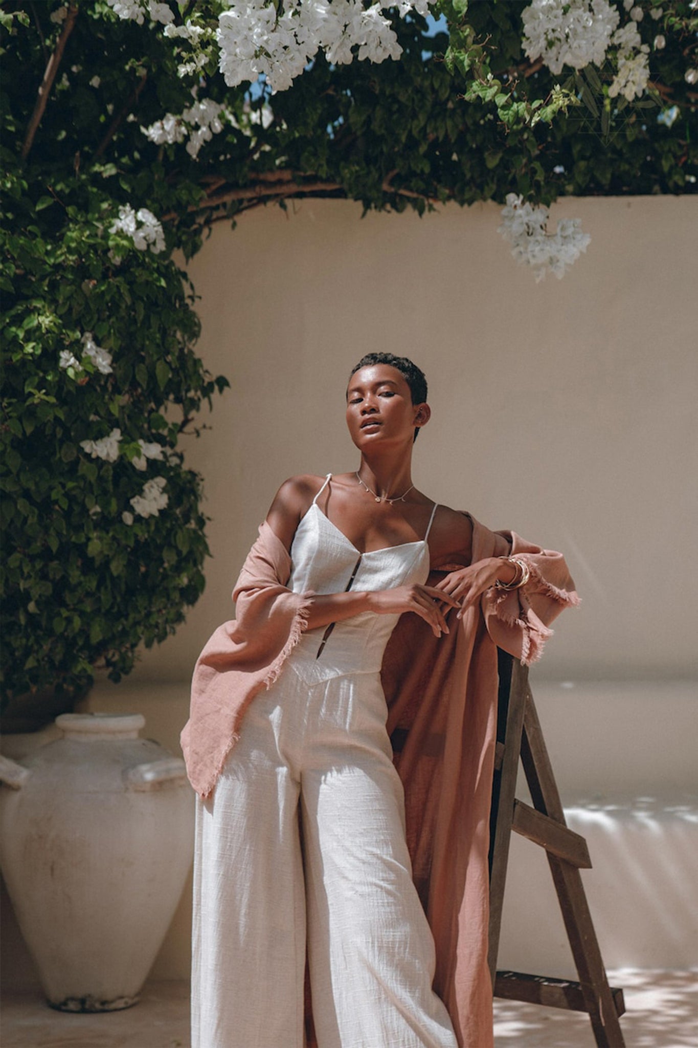 Dressed in a white outfit and a Dusty Pink Linen Cardigan, a person leans confidently against a ladder. They are outdoors, enveloped by lush greenery and delicate white flowers. Sunlight casts soft shadows, contributing to the serene and elegant ambiance. The Boho Wrap Cardigan with Belt by AYA Sacred Wear adds an extra touch of sophistication to the scene.