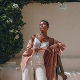 Dressed in a white outfit and a Dusty Pink Linen Cardigan, a person leans confidently against a ladder. They are outdoors, enveloped by lush greenery and delicate white flowers. Sunlight casts soft shadows, contributing to the serene and elegant ambiance. The Boho Wrap Cardigan with Belt by AYA Sacred Wear adds an extra touch of sophistication to the scene.