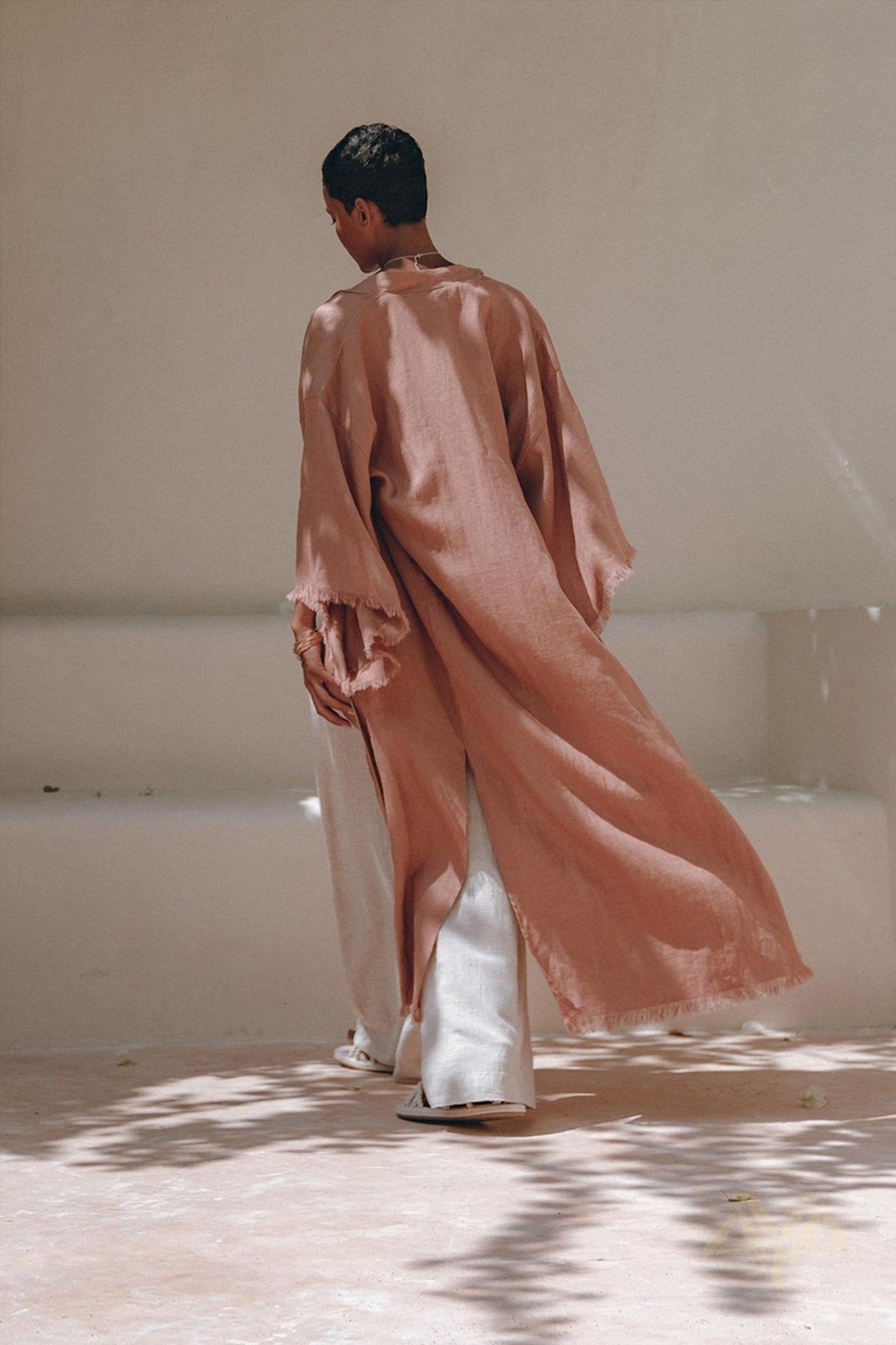 Wearing a dusty pink linen cardigan from AYA Sacred Wear, a person glides away on a light-colored path. This boho wrap overcoat, complete with its unique belt design, drapes elegantly over wide-legged cream pants. Soft shadows and light enhance the serene setting.