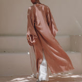 Wearing a dusty pink linen cardigan from AYA Sacred Wear, a person glides away on a light-colored path. This boho wrap overcoat, complete with its unique belt design, drapes elegantly over wide-legged cream pants. Soft shadows and light enhance the serene setting.