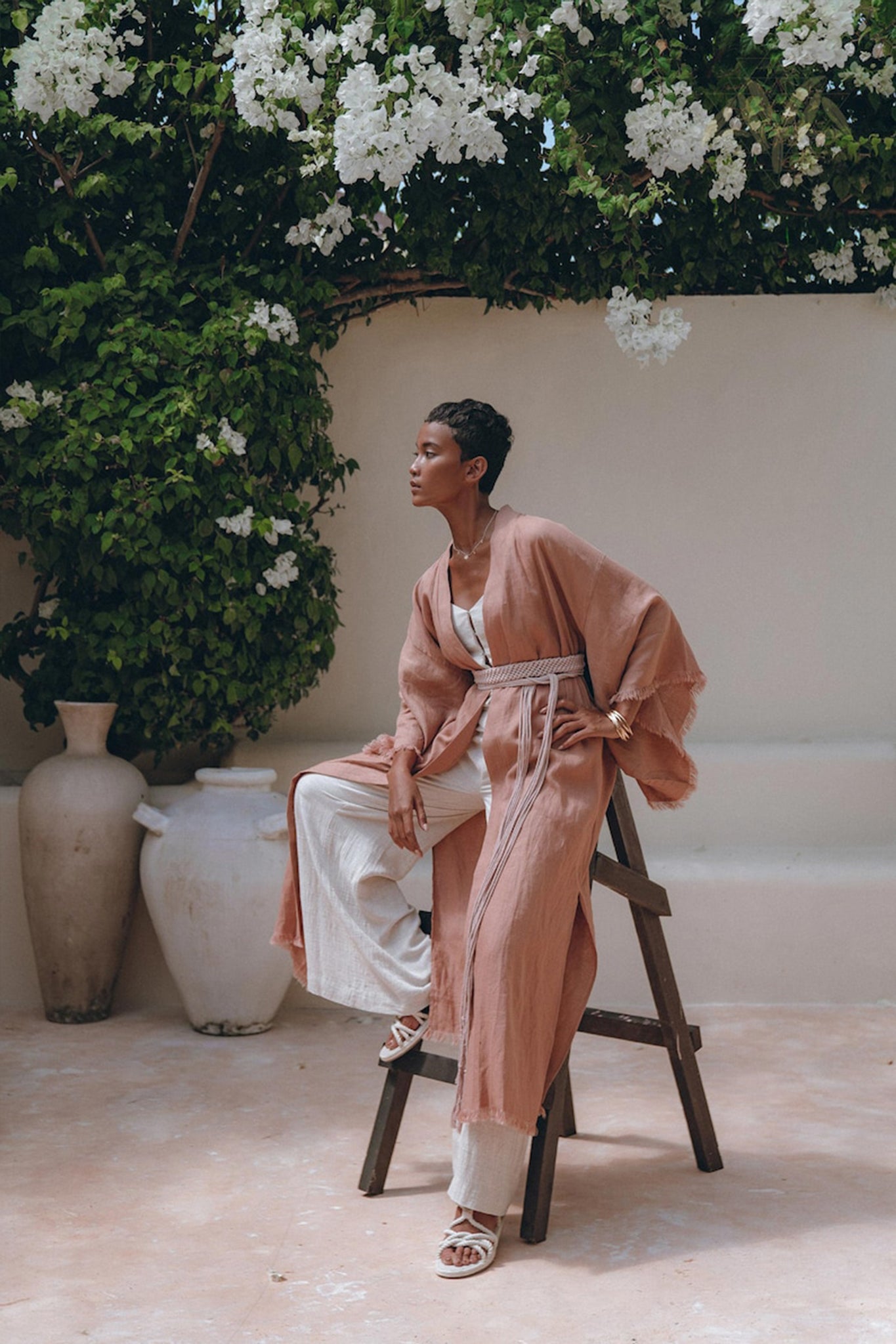 A person wearing a Dusty Pink Boho Wrap Cardigan with Belt from AYA Sacred Wear sits on a wooden stool in a bright outdoor setting. Surrounded by green foliage and white flowers, they exude tranquility. Nearby, two ceramic pots enhance the serene atmosphere with their rustic charm.