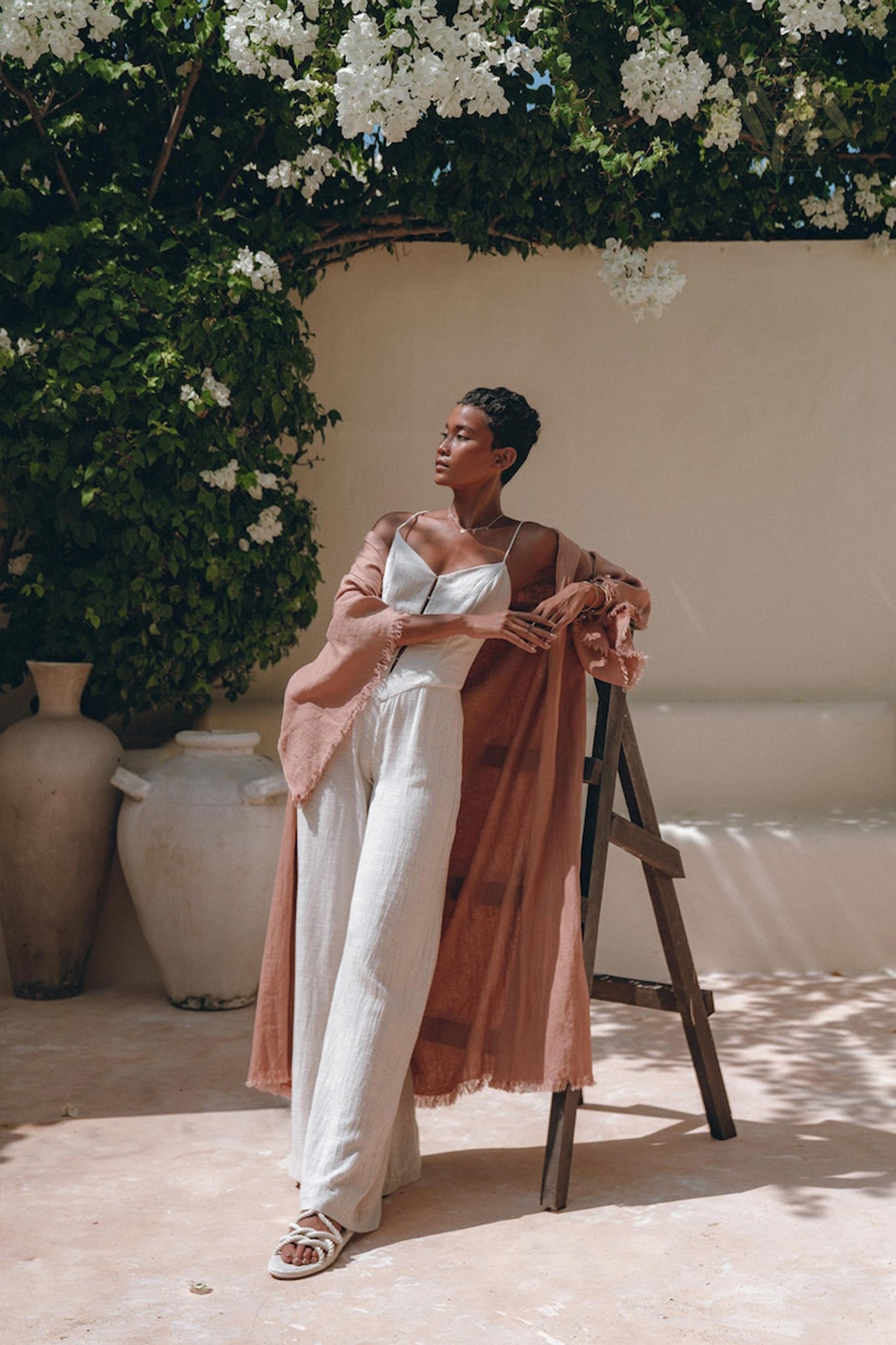 A person poses outdoors on a wooden ladder, wearing a white jumpsuit and an AYA Sacred Wear Linen Cardigan Women, a dusty pink overcoat styled as a boho wrap cardigan with a belt. They are surrounded by lush greenery, with white flowers and terracotta pots in the background.