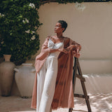A person poses outdoors on a wooden ladder, wearing a white jumpsuit and an AYA Sacred Wear Linen Cardigan Women, a dusty pink overcoat styled as a boho wrap cardigan with a belt. They are surrounded by lush greenery, with white flowers and terracotta pots in the background.