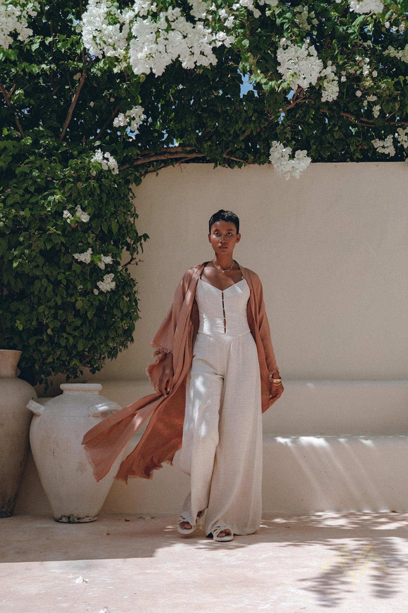 Under a canopy of white flowers, an individual stands outdoors wearing the AYA Sacred Wear Linen Cardigan, a dusty pink boho wrap with a belt, elegantly draped over a white outfit and paired with sandals. Nearby, large ceramic vases are arranged in such a way that they cast shadows on the ground.