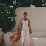 Under a canopy of white flowers, an individual stands outdoors wearing the AYA Sacred Wear Linen Cardigan, a dusty pink boho wrap with a belt, elegantly draped over a white outfit and paired with sandals. Nearby, large ceramic vases are arranged in such a way that they cast shadows on the ground.