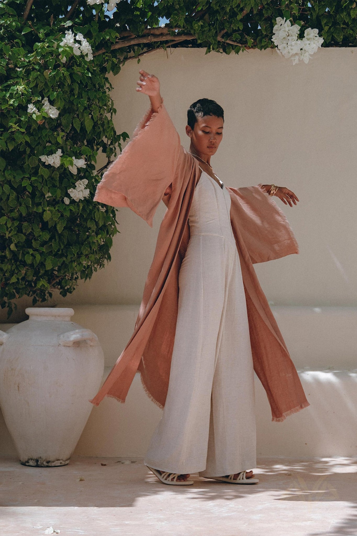 A person elegantly poses near a large ceramic pot surrounded by lush green foliage and white flowers, wearing a long, flowing dusty pink linen cardigan from AYA Sacred Wear over a white jumpsuit. The boho wrap cardigan with belt adds to the serene and stylish ambiance of the scene.