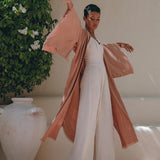 A person elegantly poses near a large ceramic pot surrounded by lush green foliage and white flowers, wearing a long, flowing dusty pink linen cardigan from AYA Sacred Wear over a white jumpsuit. The boho wrap cardigan with belt adds to the serene and stylish ambiance of the scene.