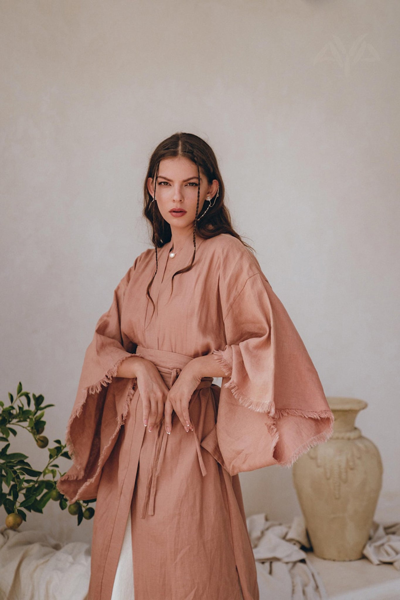 A woman with long brown hair stands in a softly lit room, wearing a flowing, belted Linen Cardigan for Women by AYA Sacred Wear in Dusty Pink. Her pensive expression adds depth to the serene scene featuring a large beige vase and lush green foliage in the background.