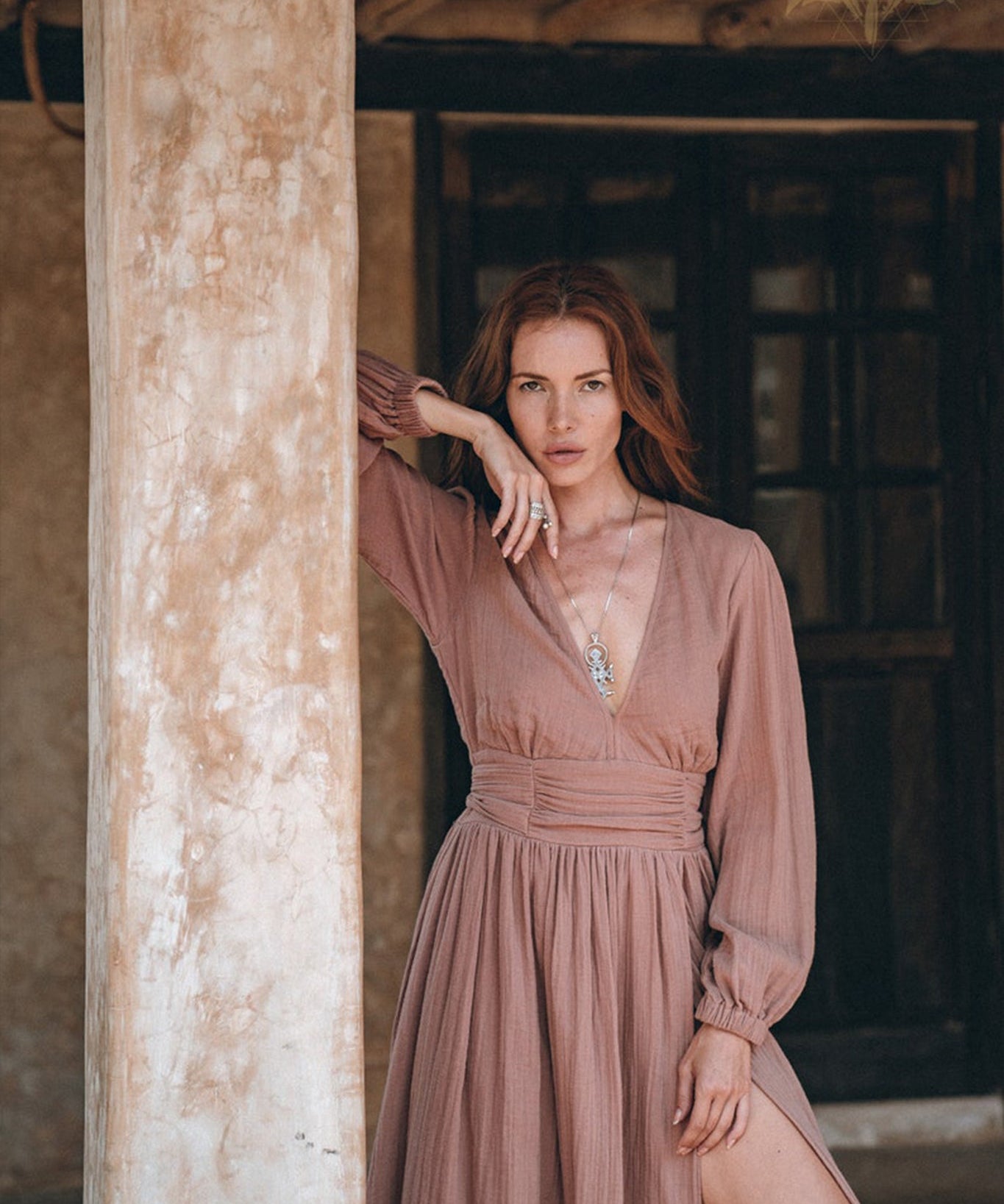 A woman with long, wavy red hair poses gracefully by a rustic wooden pillar. She is wearing AYA Sacred Wear's Long Sleeve Organic Boho Dress in taupe, featuring a deep V-neck and slit. Her right hand rests on the pillar, while her left hand touches her neck against the backdrop of a wooden door.