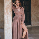 A woman with long red hair leans against a rustic pillar, showcasing the Long Sleeve Organic Boho Dress by AYA Sacred Wear. This dusty rose, bohemian bridesmaid dress features a stylish slit and is paired with brown ankle boots. She strikes a pose, one arm on the pillar and her hand gently touching her head, set against an earthy and textured backdrop.