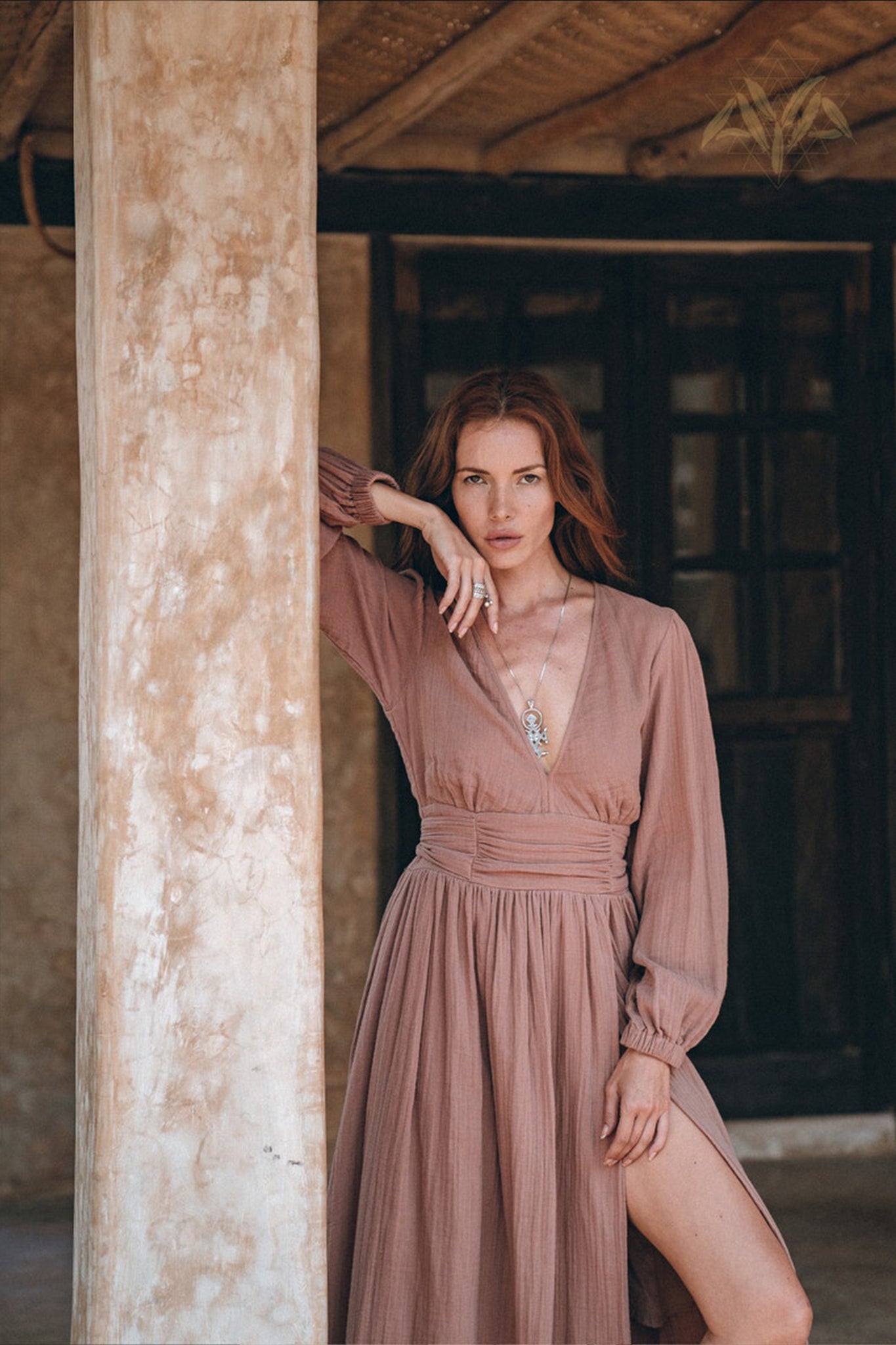 A woman with long, wavy red hair poses gracefully by a rustic wooden pillar. She is wearing AYA Sacred Wear's Long Sleeve Organic Boho Dress in taupe, featuring a deep V-neck and slit. Her right hand rests on the pillar, while her left hand touches her neck against the backdrop of a wooden door.