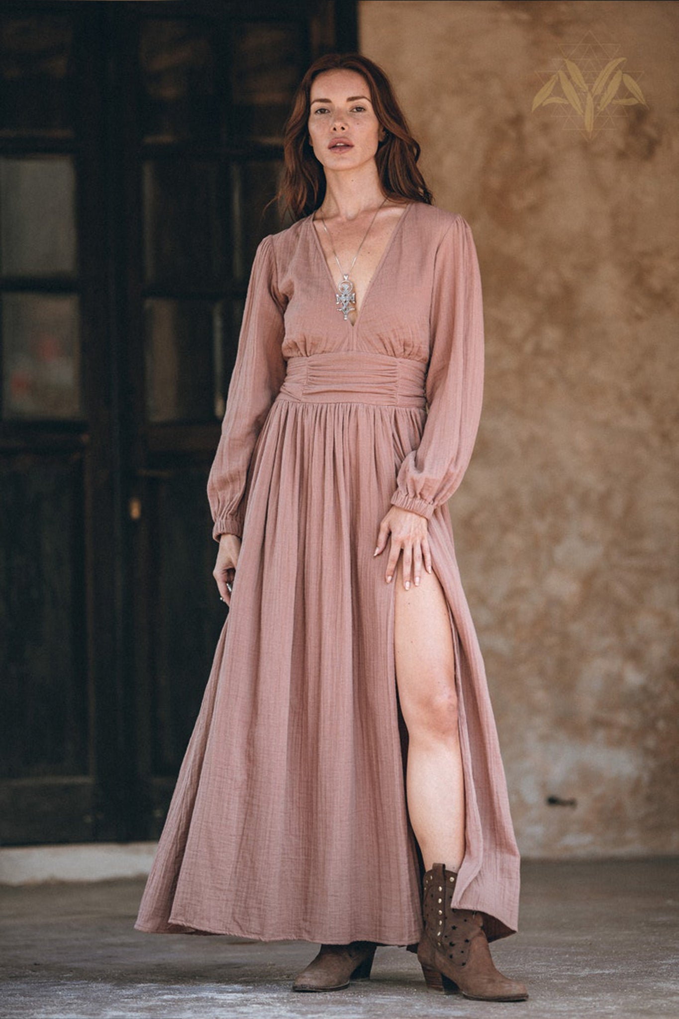 In front of a rustic background, a woman embodies an autumn boho vibe in the Long Sleeve Organic Boho Dress by AYA Sacred Wear. The dusty rose dress features a deep slit and is paired with a necklace and ankle boots, as she holds an unspecified object in her left hand.