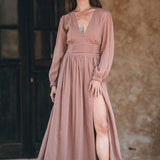 In front of a rustic background, a woman embodies an autumn boho vibe in the Long Sleeve Organic Boho Dress by AYA Sacred Wear. The dusty rose dress features a deep slit and is paired with a necklace and ankle boots, as she holds an unspecified object in her left hand.