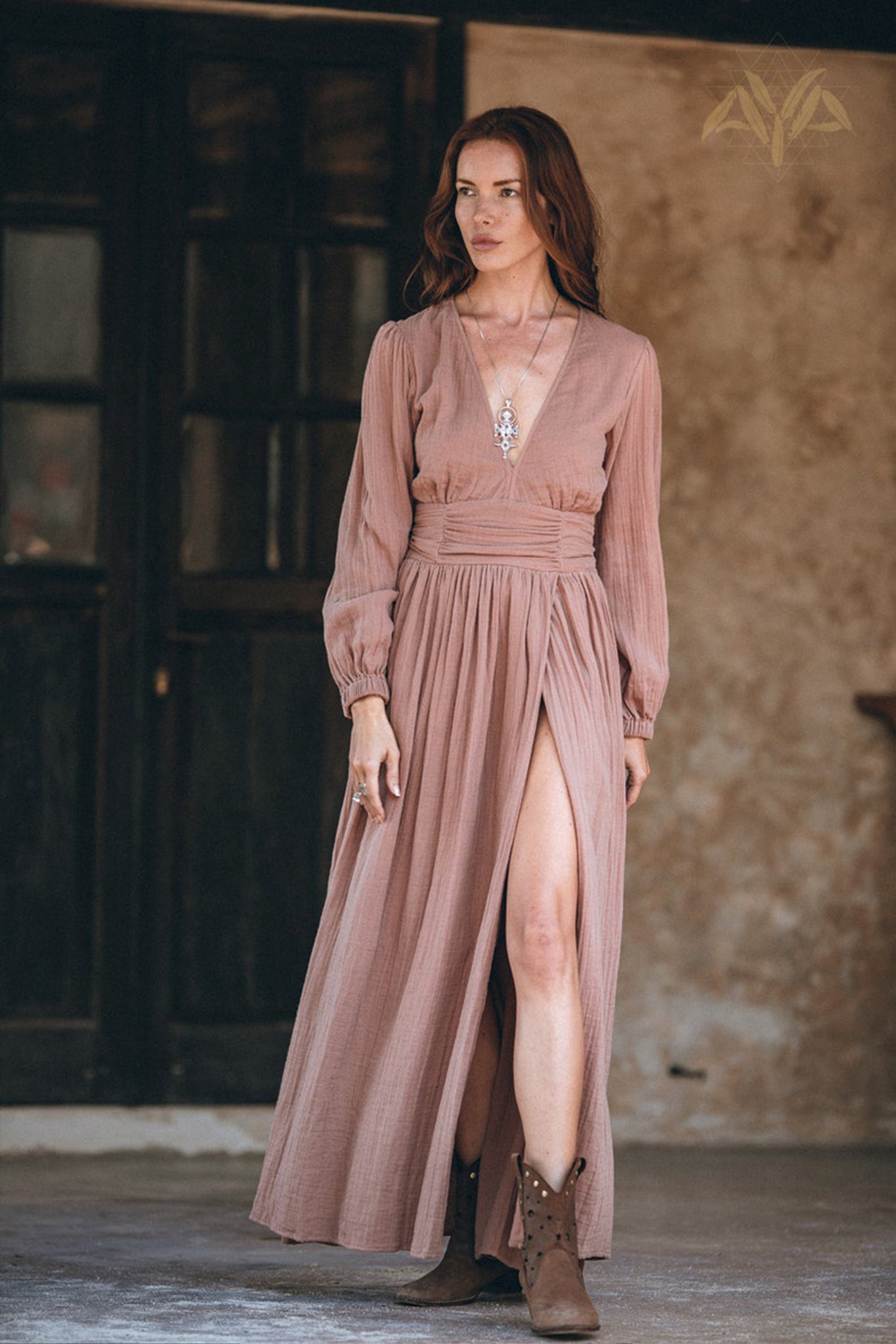 A woman stands in front of a rustic building wearing the Long Sleeve Organic Boho Dress by AYA Sacred Wear, which features a high slit and long sleeves. She has long hair, is accessorized with a necklace, and finishes her look with brown ankle boots.