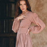 A woman with long brown hair dons the Long Sleeve Organic Boho Dress by AYA Sacred Wear, a flowing autumn bohemian dress in dusty pink, as she stands against a textured wall. Her left hand, adorned with multiple rings, gently touches her face, complementing the warm and rustic setting.