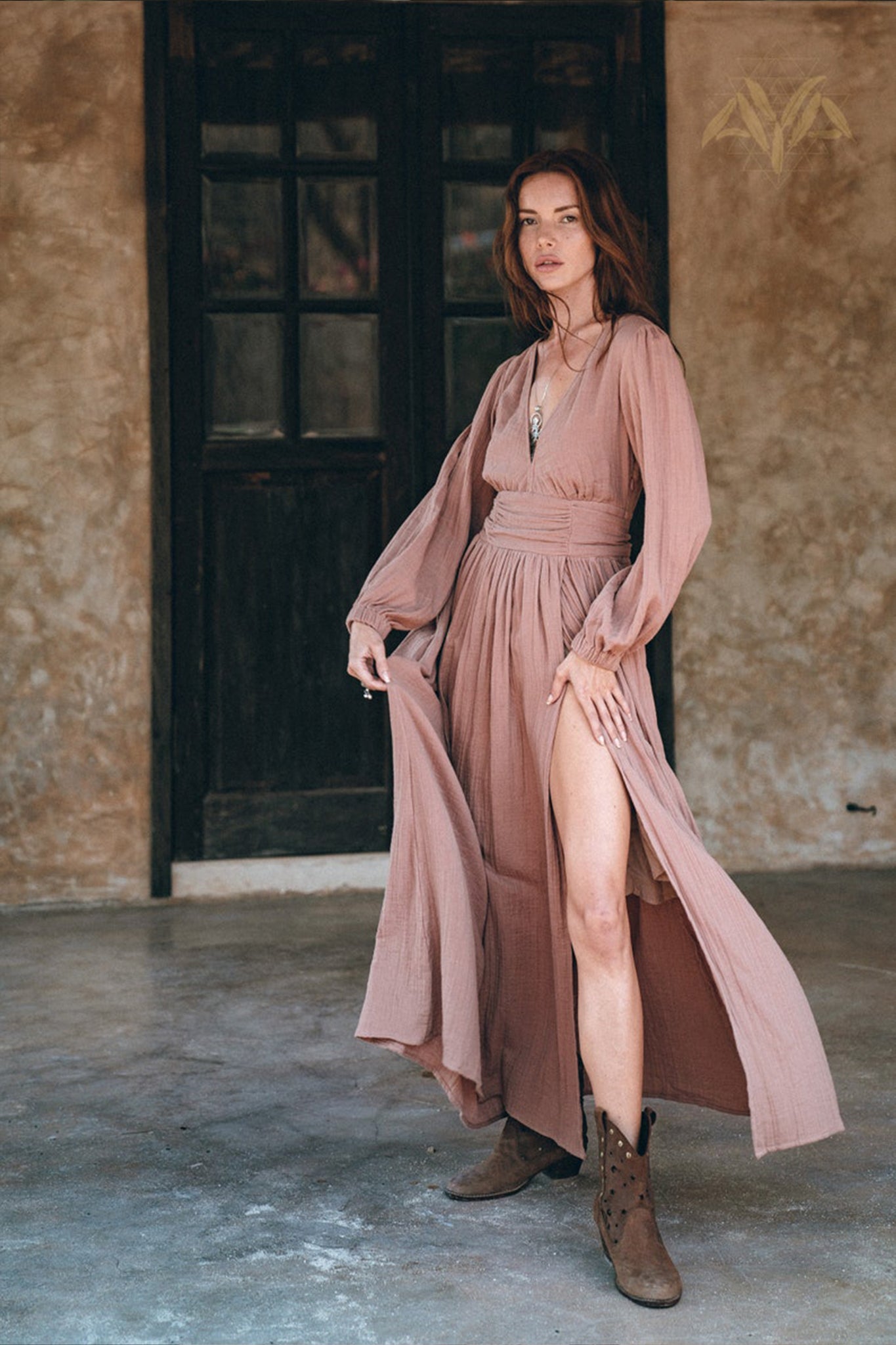 A woman poses indoors wearing the Long Sleeve Organic Boho Dress by AYA Sacred Wear. This dusty rose maxi dress features long sleeves and a high slit, styled with a necklace and brown ankle boots. She stands on a concrete floor, set against the backdrop of a dark wooden door and a textured wall.