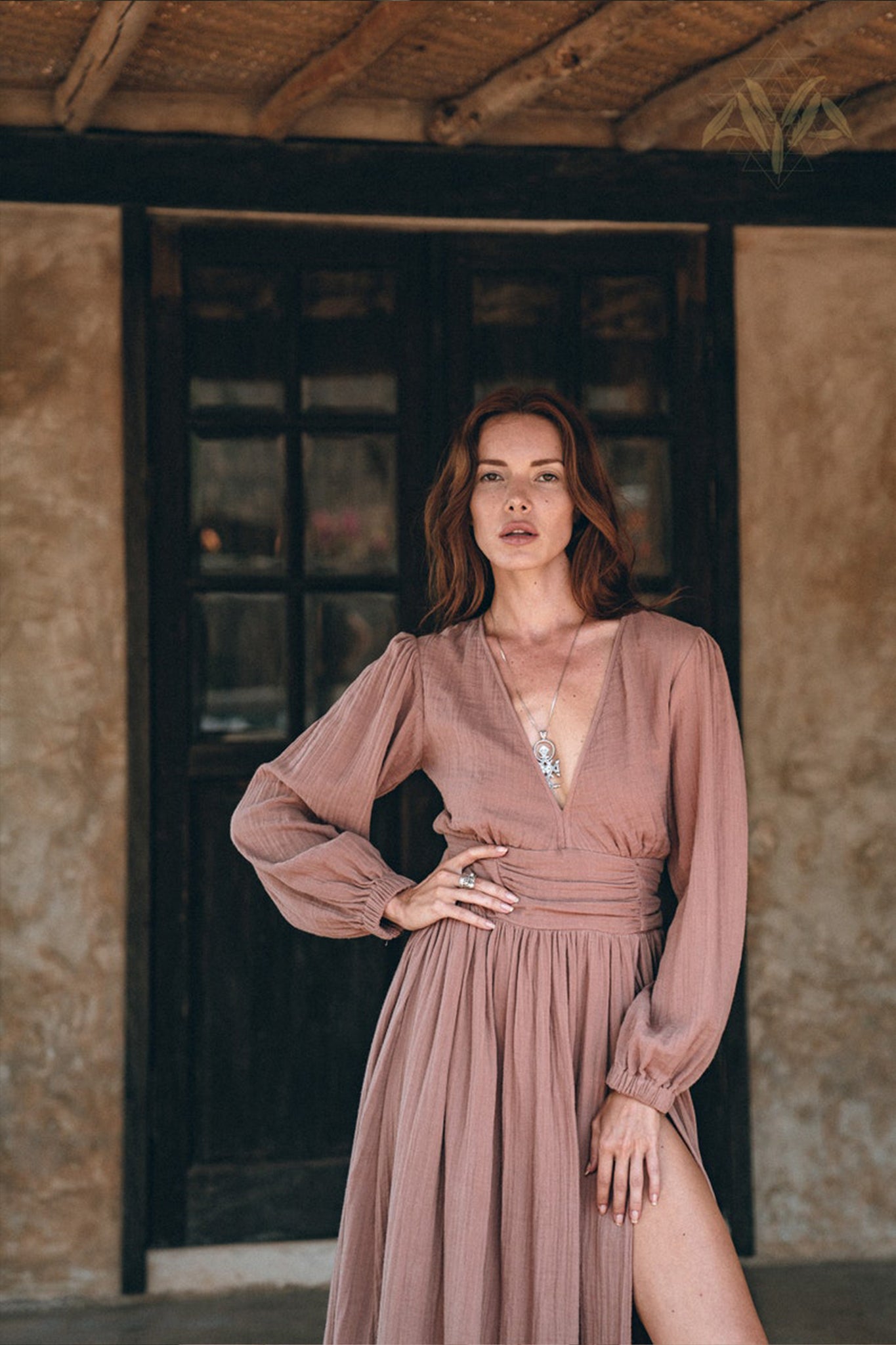 A woman with long hair stands confidently in front of a rustic wooden door, wearing the Long Sleeve Organic Boho Dress by AYA Sacred Wear. The dress, in a dusty rose hue with a deep V-neck, perfectly embodies boho elegance. One hand rests on her hip as she poses against a backdrop of textured walls and wooden beam ceilings, enhancing the scene's charm.