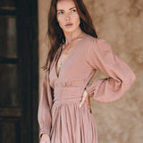 A woman with long brown hair is wearing a flowing Long Sleeve Organic Boho Dress by AYA Sacred Wear, suitable for bridesmaids. She poses with one hand on her hip and the other holding the dress. The background is a neutral brown, featuring a rustic wooden door.