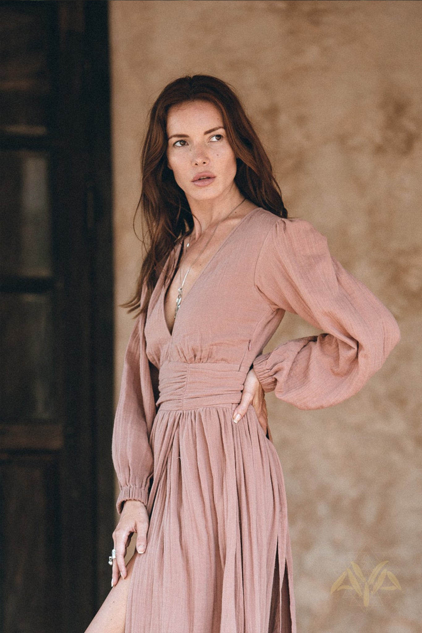 A woman with long brown hair is wearing a flowing Long Sleeve Organic Boho Dress by AYA Sacred Wear, suitable for bridesmaids. She poses with one hand on her hip and the other holding the dress. The background is a neutral brown, featuring a rustic wooden door.