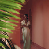 A person in a long, flowing Macramé Belted Boho Dress by AYA Sacred Wear, adorned with handmade ombre organic dyes, stands in a pink-walled corridor. Green leaves frame the scene to the left, creating a serene and stylish atmosphere enhanced by soft lighting.