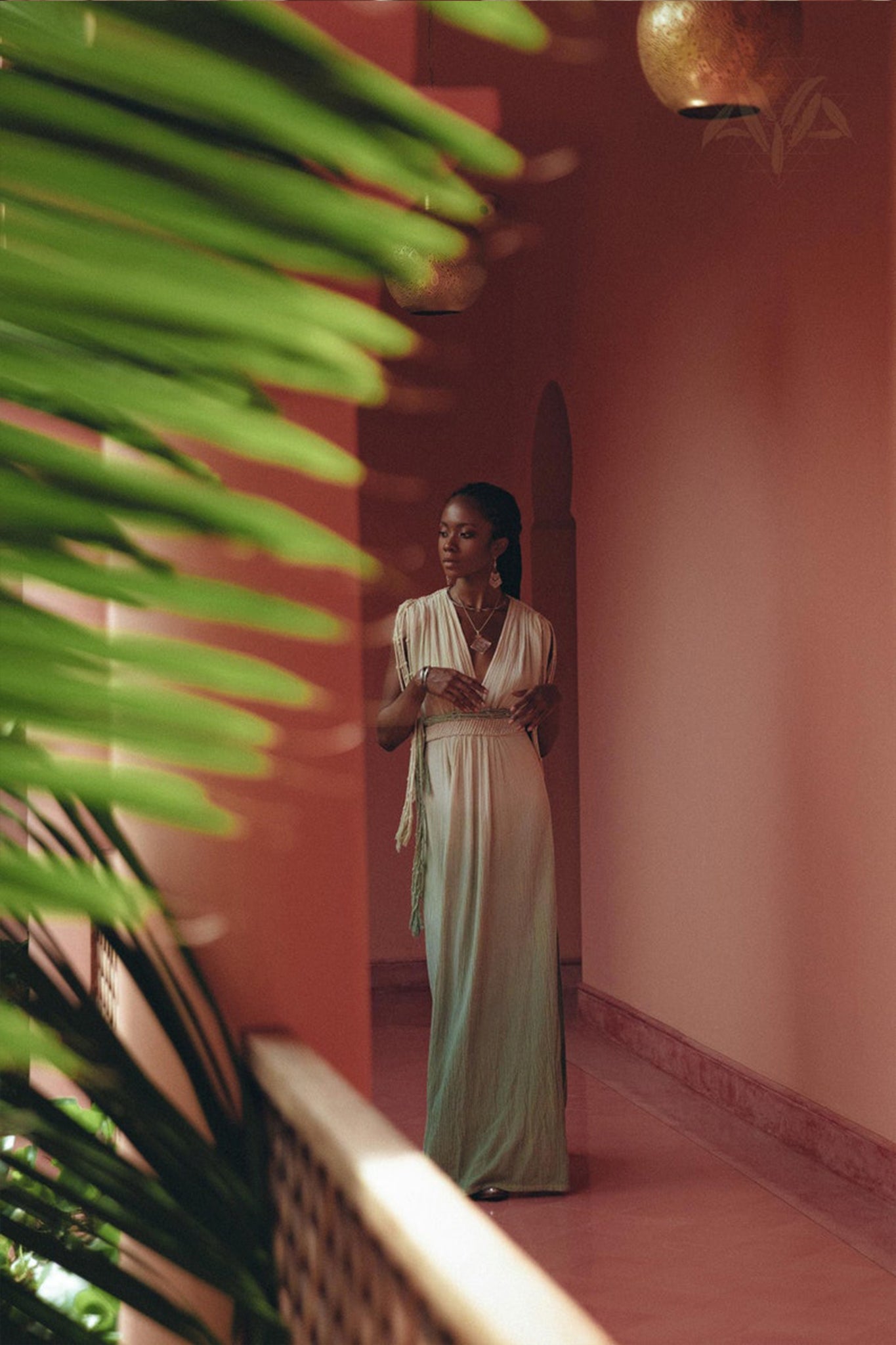 A person in a long, flowing Macramé Belted Boho Dress by AYA Sacred Wear, adorned with handmade ombre organic dyes, stands in a pink-walled corridor. Green leaves frame the scene to the left, creating a serene and stylish atmosphere enhanced by soft lighting.