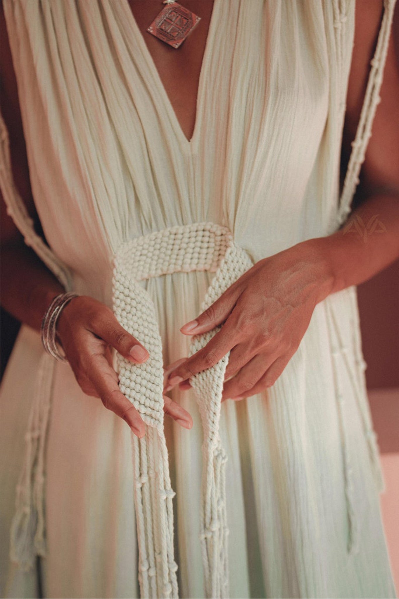 Dressed in the Macramé Belted Boho Dress by AYA Sacred Wear, which is a handmade ombre organically dyed dress, a person holds the woven belt tied around their waist. Silver bracelets adorn their hands, and the dress showcases a deep V-neck with delicate fringe details.