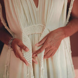 Dressed in the Macramé Belted Boho Dress by AYA Sacred Wear, which is a handmade ombre organically dyed dress, a person holds the woven belt tied around their waist. Silver bracelets adorn their hands, and the dress showcases a deep V-neck with delicate fringe details.