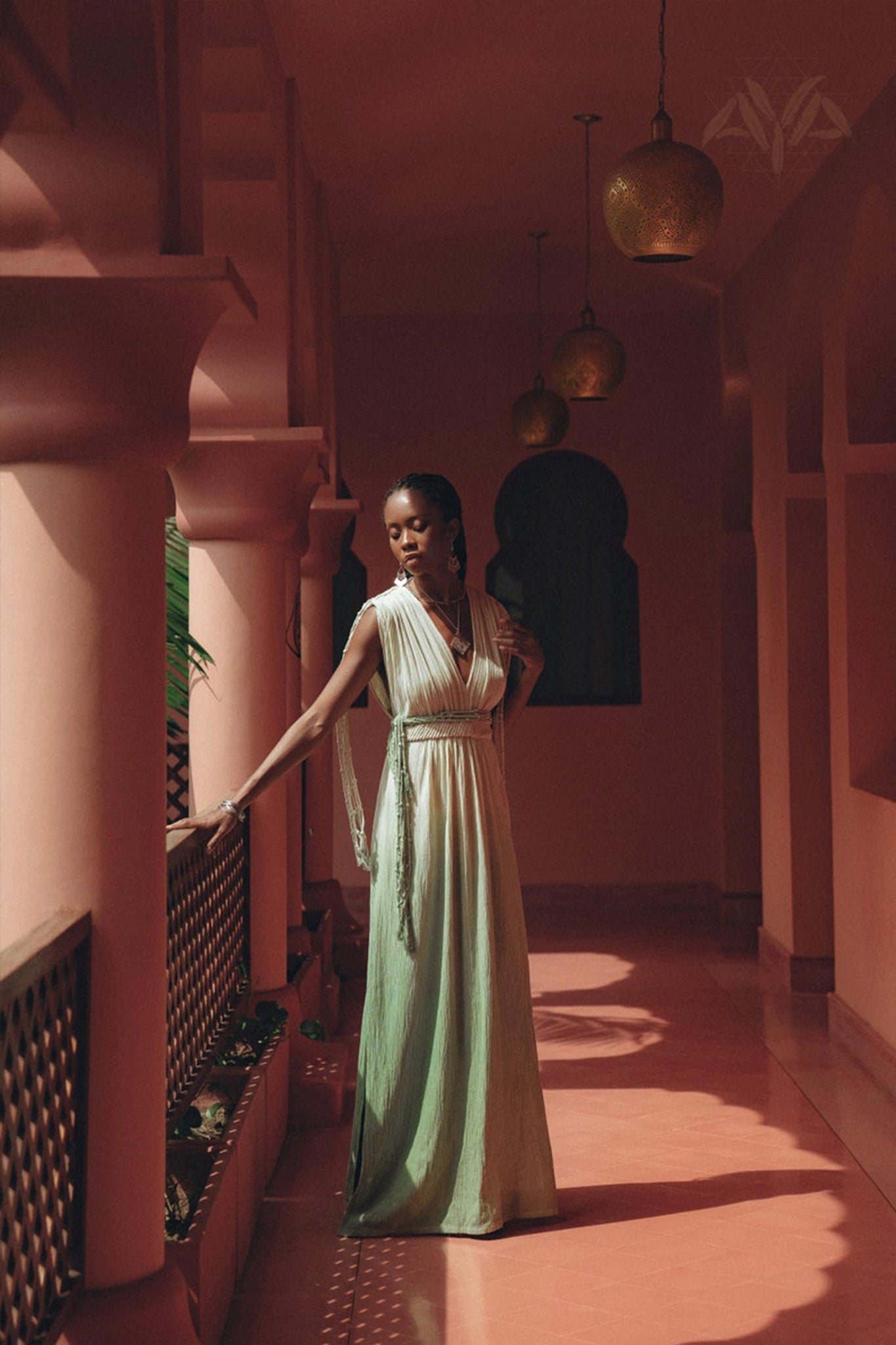 A woman wearing an AYA Sacred Wear Macramé Belted Boho Dress, handcrafted and organically ombre dyed, stands in a sunlit corridor adorned with decorative arches and lanterns. She gazes thoughtfully to the side as intricate railing shadows cast patterns on the floor.