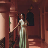 A woman wearing an AYA Sacred Wear Macramé Belted Boho Dress, handcrafted and organically ombre dyed, stands in a sunlit corridor adorned with decorative arches and lanterns. She gazes thoughtfully to the side as intricate railing shadows cast patterns on the floor.