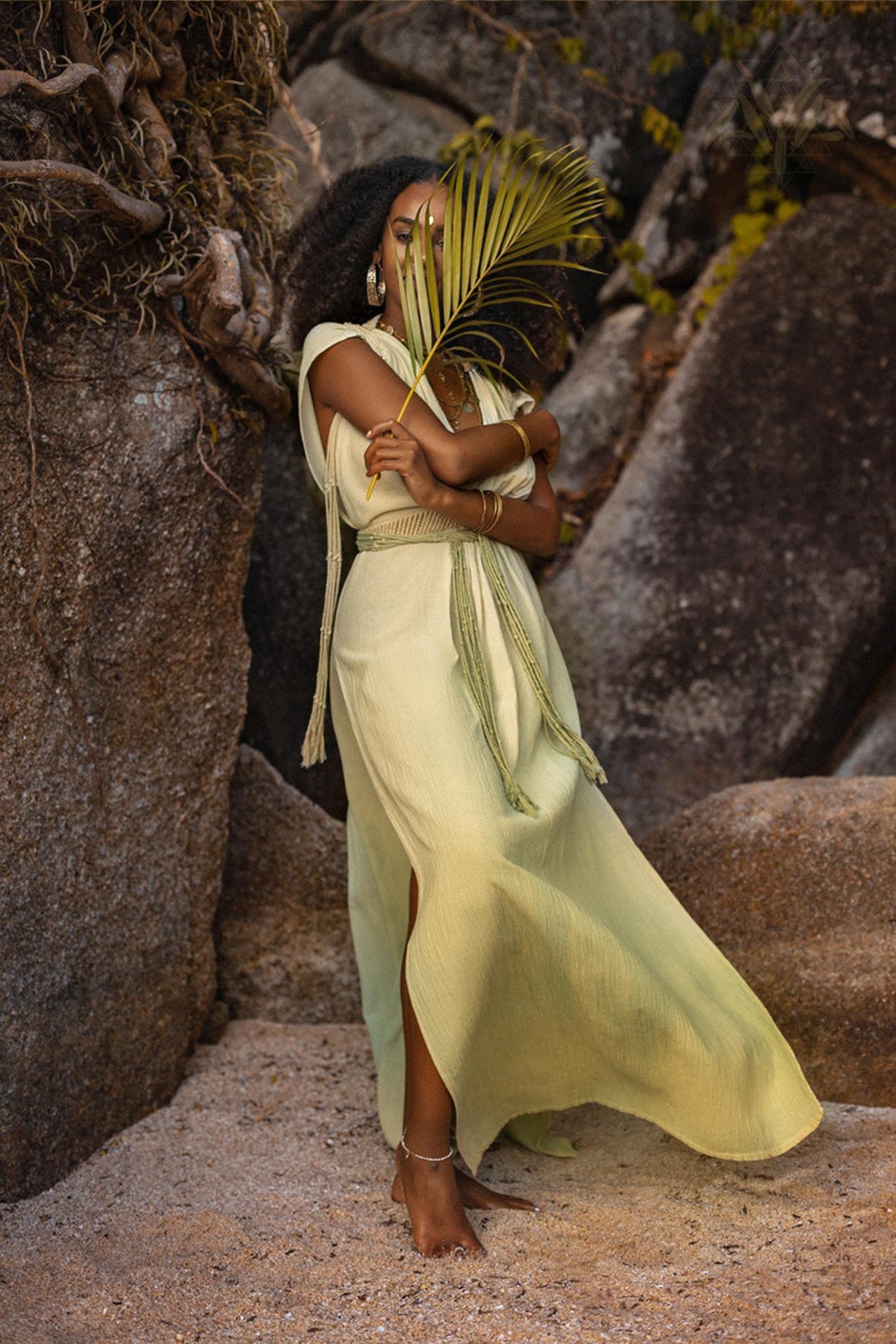 A woman wearing the Macramé Belted Boho Dress, a handmade ombre organically dyed creation by AYA Sacred Wear, stands on a sandy beach beside large rocks. She holds a palm leaf covering part of her face. Her curly hair cascades down as she wears gold jewelry, including earrings and bracelets, creating a serene and natural scene.