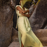 A woman wearing the Macramé Belted Boho Dress, a handmade ombre organically dyed creation by AYA Sacred Wear, stands on a sandy beach beside large rocks. She holds a palm leaf covering part of her face. Her curly hair cascades down as she wears gold jewelry, including earrings and bracelets, creating a serene and natural scene.