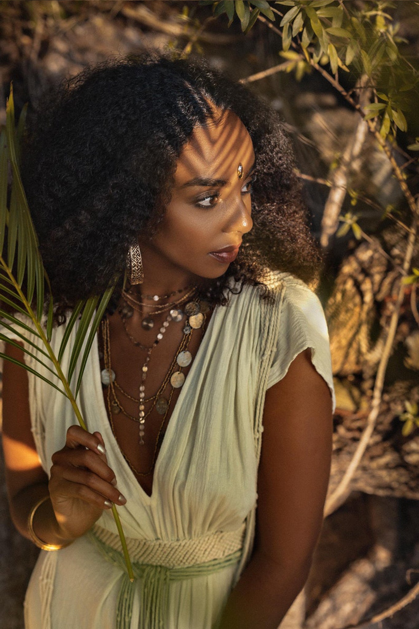 A woman with curly hair stands under the play of dappled light, holding a palm leaf. She is adorned in a Greek-style Macramé Belted Boho Dress by AYA Sacred Wear, featuring a handmade ombre organic dye. Layered necklaces and earrings complete her look as she gazes to her left, surrounded by a serene backdrop of foliage and rocks.