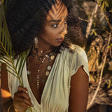 A woman with curly hair stands under the play of dappled light, holding a palm leaf. She is adorned in a Greek-style Macramé Belted Boho Dress by AYA Sacred Wear, featuring a handmade ombre organic dye. Layered necklaces and earrings complete her look as she gazes to her left, surrounded by a serene backdrop of foliage and rocks.