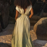 A woman with long curly hair stands on a rocky beach, her Macramé Belted Boho Dress from AYA Sacred Wear flowing gracefully with an open back. She faces away from the camera, embraced by natural rock formations and sandy ground.