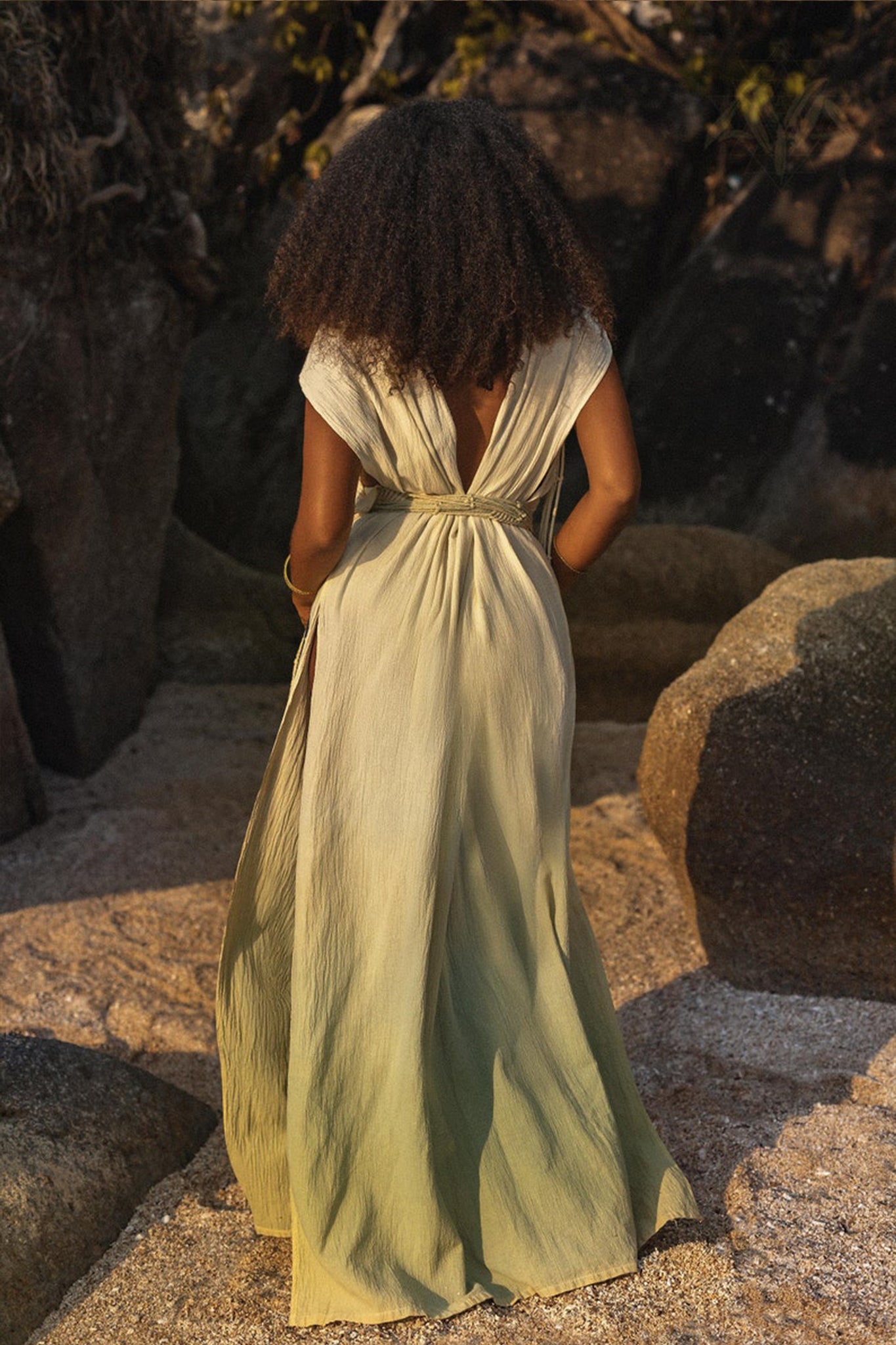 A woman with long curly hair stands on a rocky beach, her Macramé Belted Boho Dress from AYA Sacred Wear flowing gracefully with an open back. She faces away from the camera, embraced by natural rock formations and sandy ground.