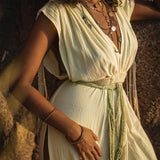 A woman with curly hair wears the Macramé Belted Boho Dress, a handmade ombre organically dyed dress by AYA Sacred Wear, featuring a flowing Greek style with a slit. Layered necklaces and bracelets adorn her as she leans against a rock wall in soft sunlight, creating a warm and serene ambiance.