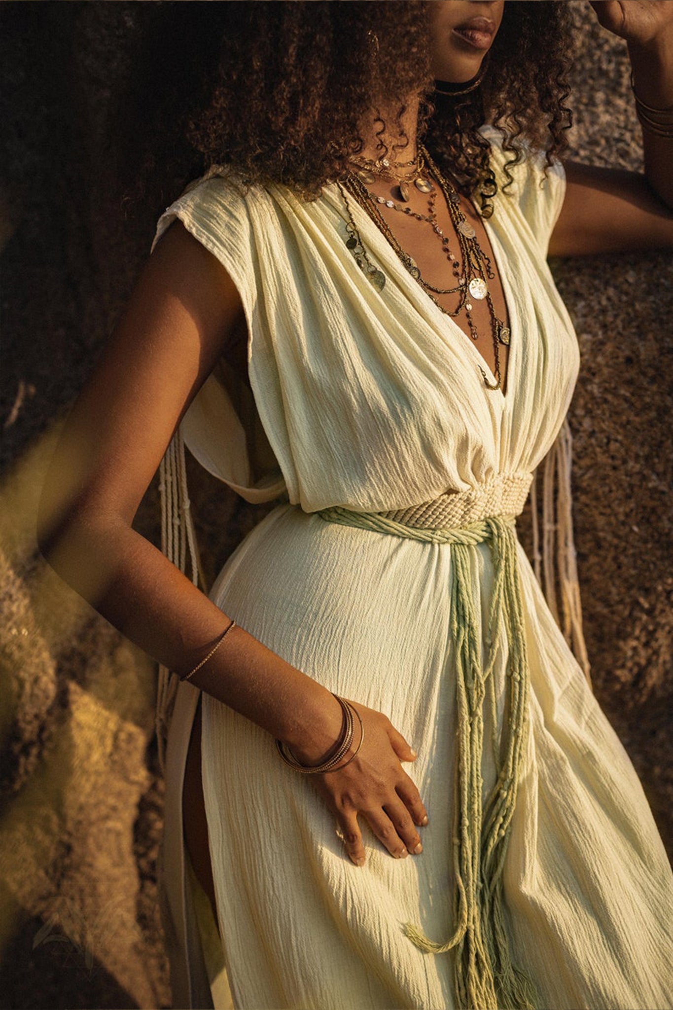 A woman with curly hair wears the Macramé Belted Boho Dress, a handmade ombre organically dyed dress by AYA Sacred Wear, featuring a flowing Greek style with a slit. Layered necklaces and bracelets adorn her as she leans against a rock wall in soft sunlight, creating a warm and serene ambiance.