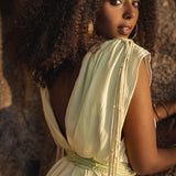 Dressed in a Macramé Belted Boho Dress from AYA Sacred Wear, the woman with curly hair complements her Greek-inspired attire with gold jewelry and a delicate gold ornament adorning her forehead. She stands against a rocky backdrop, glancing over her shoulder in the warm glow.