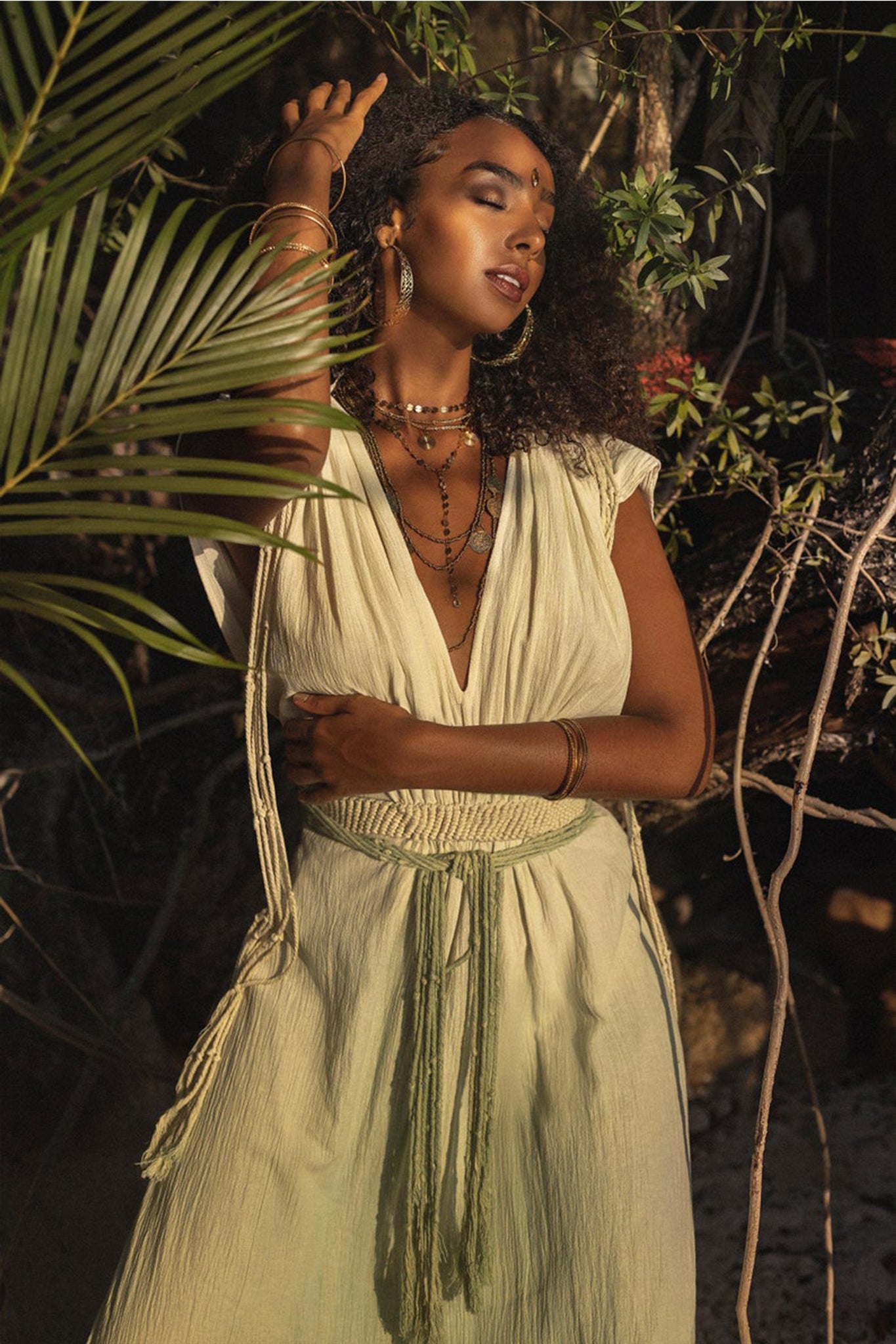 A woman wearing the Macramé Belted Boho Dress, a handmade ombre organically dyed dress by AYA Sacred Wear, stands amid lush greenery. She complements her look with layered necklaces and bracelets. Her eyes are closed as she gently touches her braided hair, savoring the sunlight filtering through the trees.