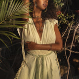 A woman wearing the Macramé Belted Boho Dress, a handmade ombre organically dyed dress by AYA Sacred Wear, stands amid lush greenery. She complements her look with layered necklaces and bracelets. Her eyes are closed as she gently touches her braided hair, savoring the sunlight filtering through the trees.