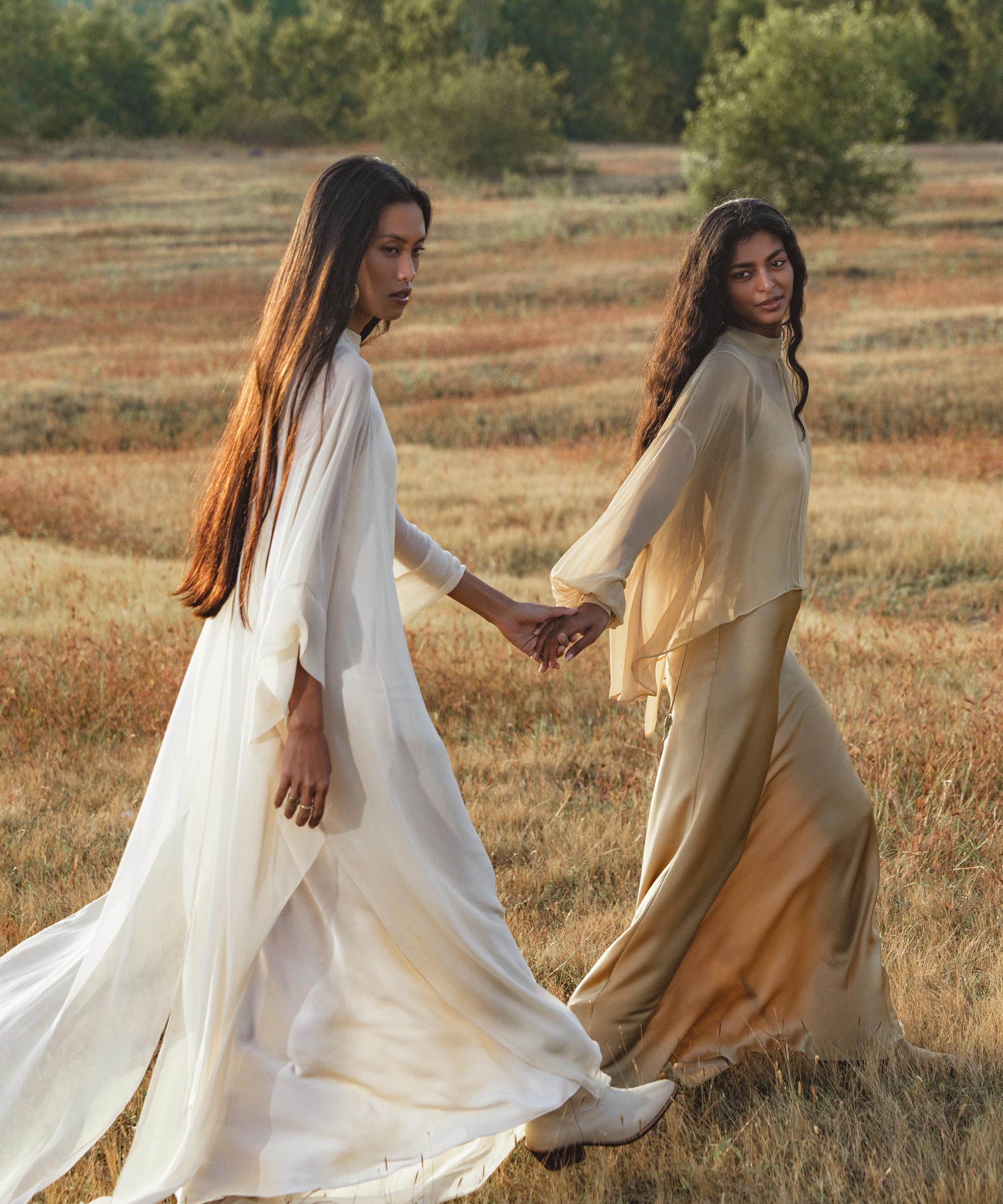 Two women with long hair walk hand-in-hand through a grassy field. Both wear AYA Sacred Wear Mahadevi Dress Cream—pure silk chiffon goddess gowns—in white and beige, framed by trees and a cloudy sky for a serene, natural vibe.