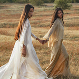 Two women with long brown hair walk hand in hand through a meadow. One wears the Mahadevi Dress Cream – Pure Silk Chiffon Goddess Gown by AYA Sacred Wear, while the other wears a loose beige dress. Golden grass and green trees frame their stroll.
