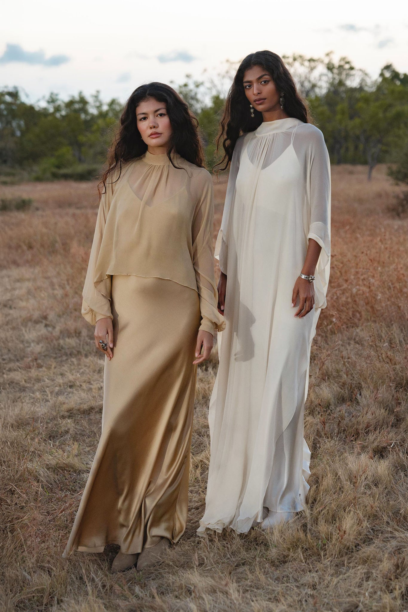 Two women stand in a dry, grassy field. One wears the AYA Sacred Wear Mahadevi Dress Cream—a pure silk chiffon goddess gown—while the other dons a handcrafted flowing dress. Their long dark hair frames serene faces, trees and blue sky behind them.