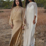 Two women stand in a dry, grassy field. One wears the AYA Sacred Wear Mahadevi Dress Cream—a pure silk chiffon goddess gown—while the other dons a handcrafted flowing dress. Their long dark hair frames serene faces, trees and blue sky behind them.