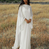 A woman with long, wavy dark hair stands in a sunlit grassy field wearing the AYA Sacred Wear Mahadevi Dress Cream—an elegant pure silk chiffon goddess gown—against a serene backdrop of hills and blue sky, gazing softly ahead with hands gently clasped.